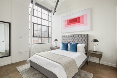 Modern loft bedroom with gray upholstered headboard, pink contemporary art, concrete floors, rug, nightstand, blue pillows, large industrial window.